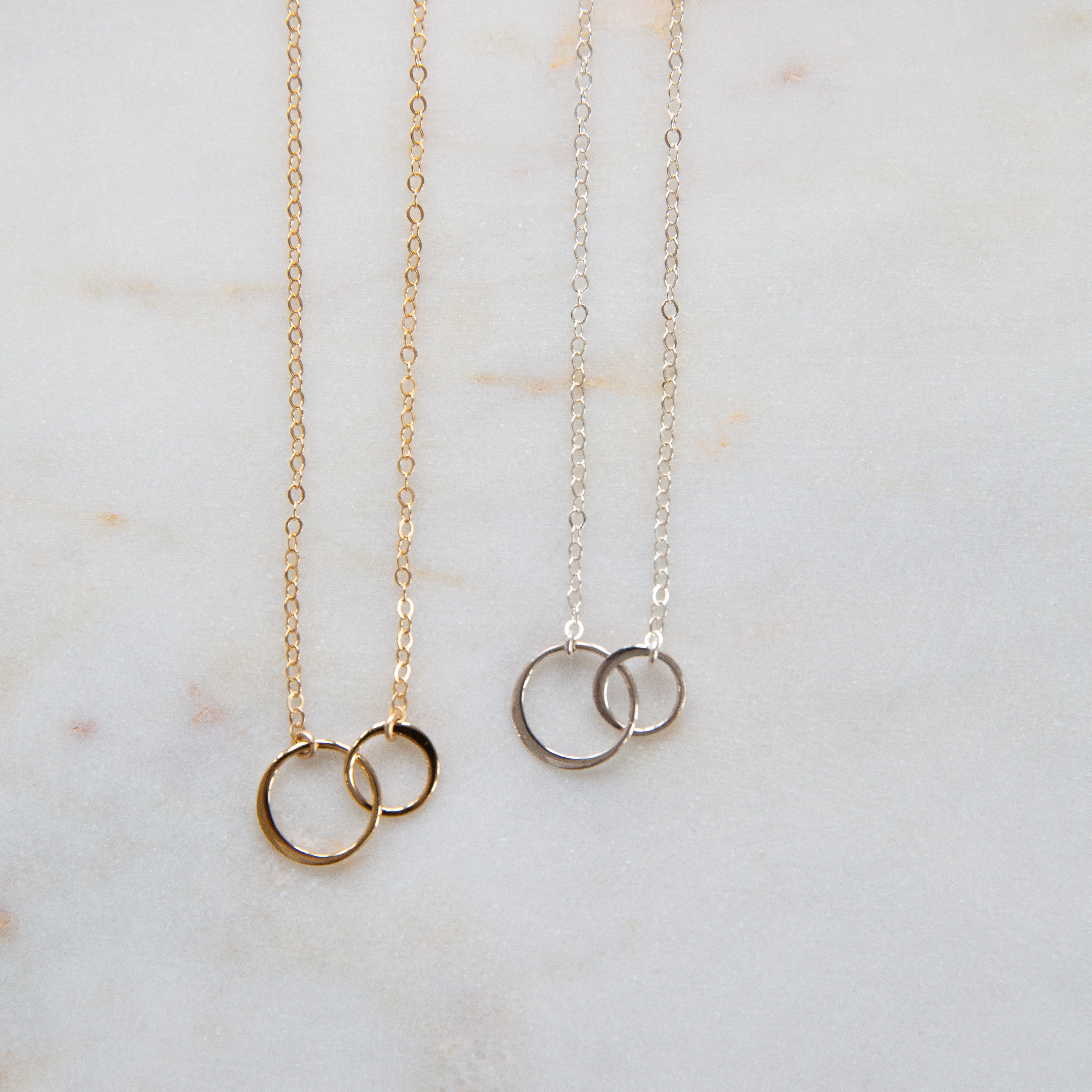 Two necklaces, one with gold chains and circles, the other with silver chains and circles, displayed on a marble surface.