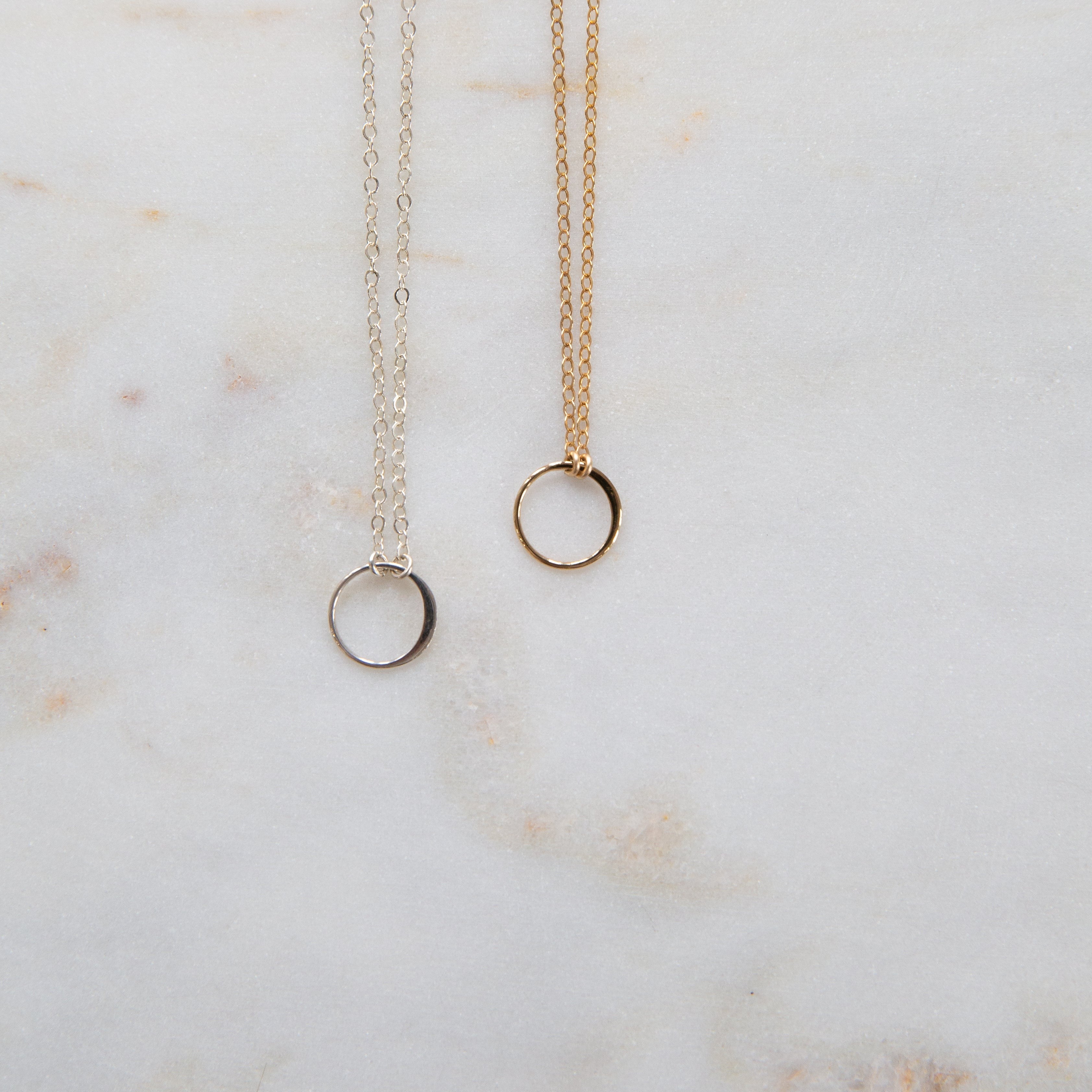 Two necklaces, one with a silver circle pendant and the other with a gold circle pendant, laid out on a marble surface.