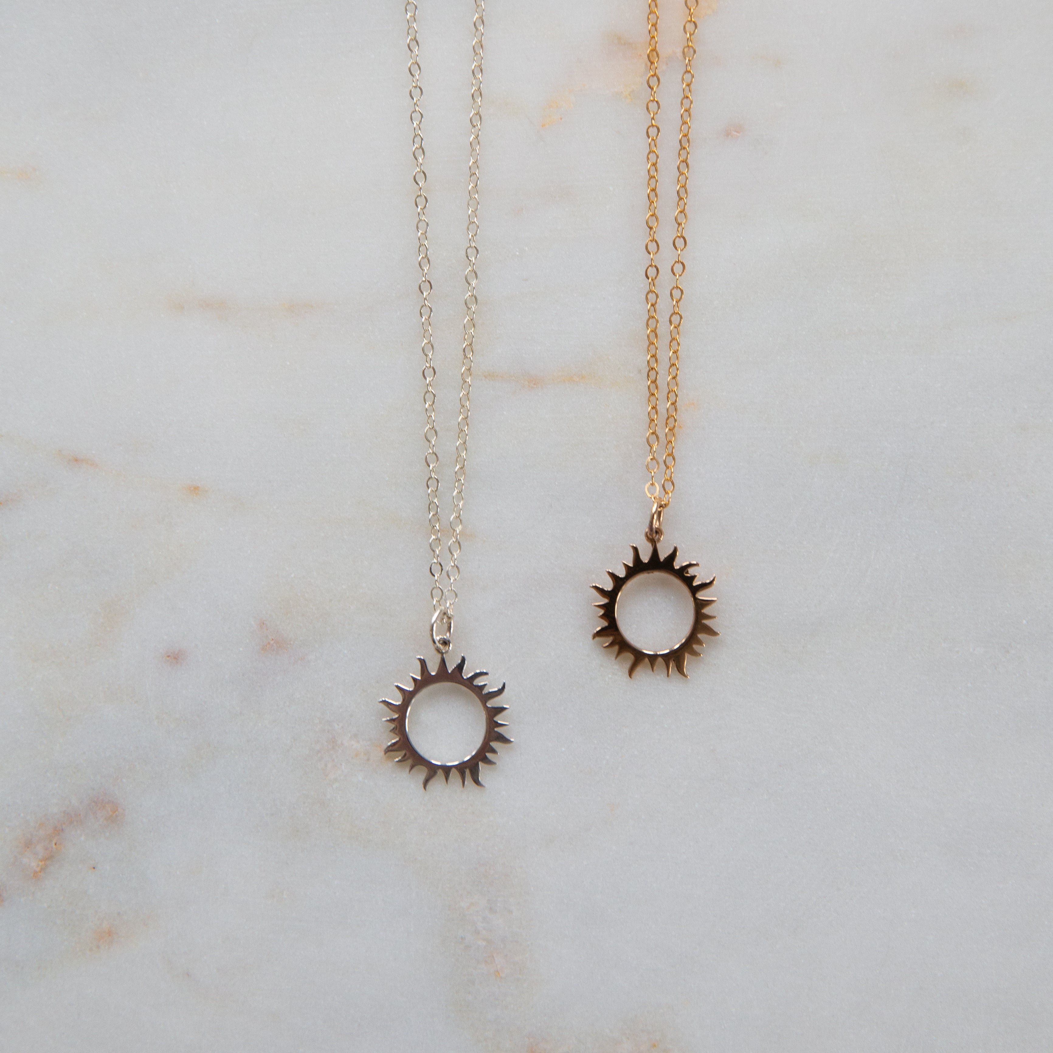 Two necklaces, one with a silver sun pendant and the other with a gold sun pendant, laid out on a marble surface.
