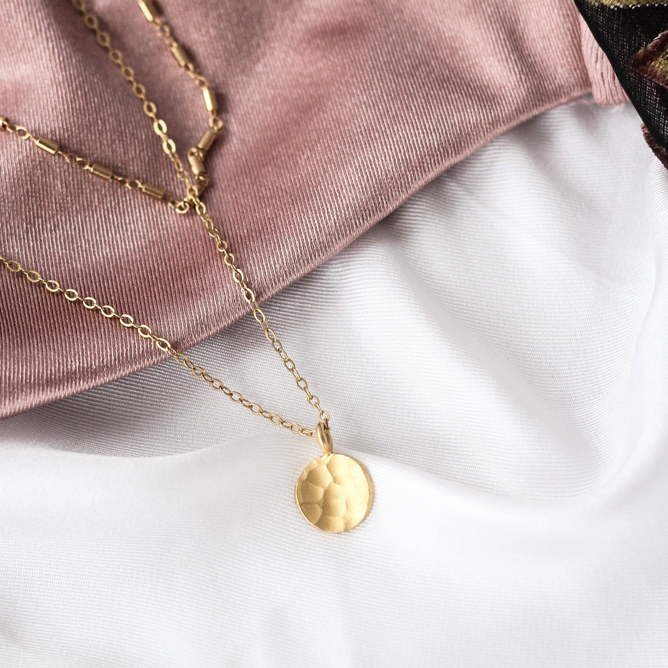 A gold-colored hammered dot necklace laid out on a flat surface with a pink fabric in the background.