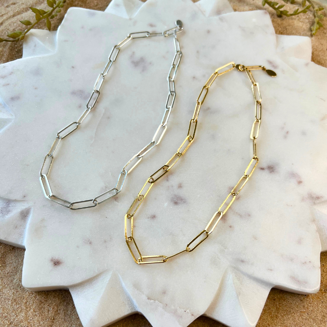 Two necklaces, one in silver and one in gold, laid on a marble surface. They have a bold, link chain design.