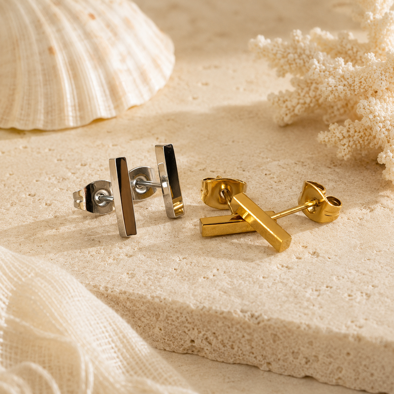 Gold and silver earrings on a sandy surface with a shell and coral in the background