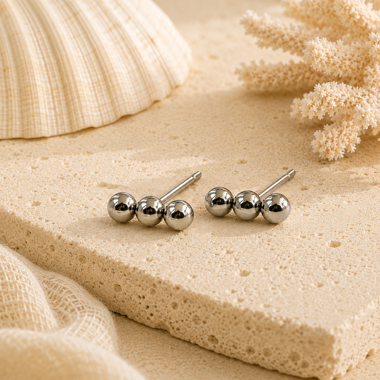 Silver ball earrings on a sandy surface with shells and coral in the background