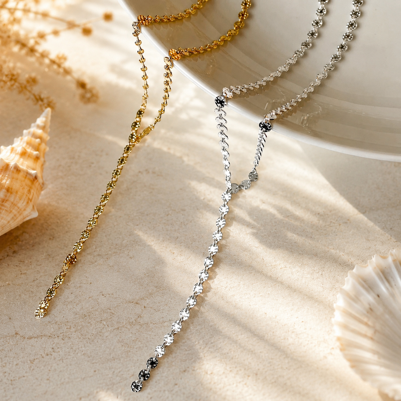 Two necklaces, one gold and one silver, on a sandy surface with shells.