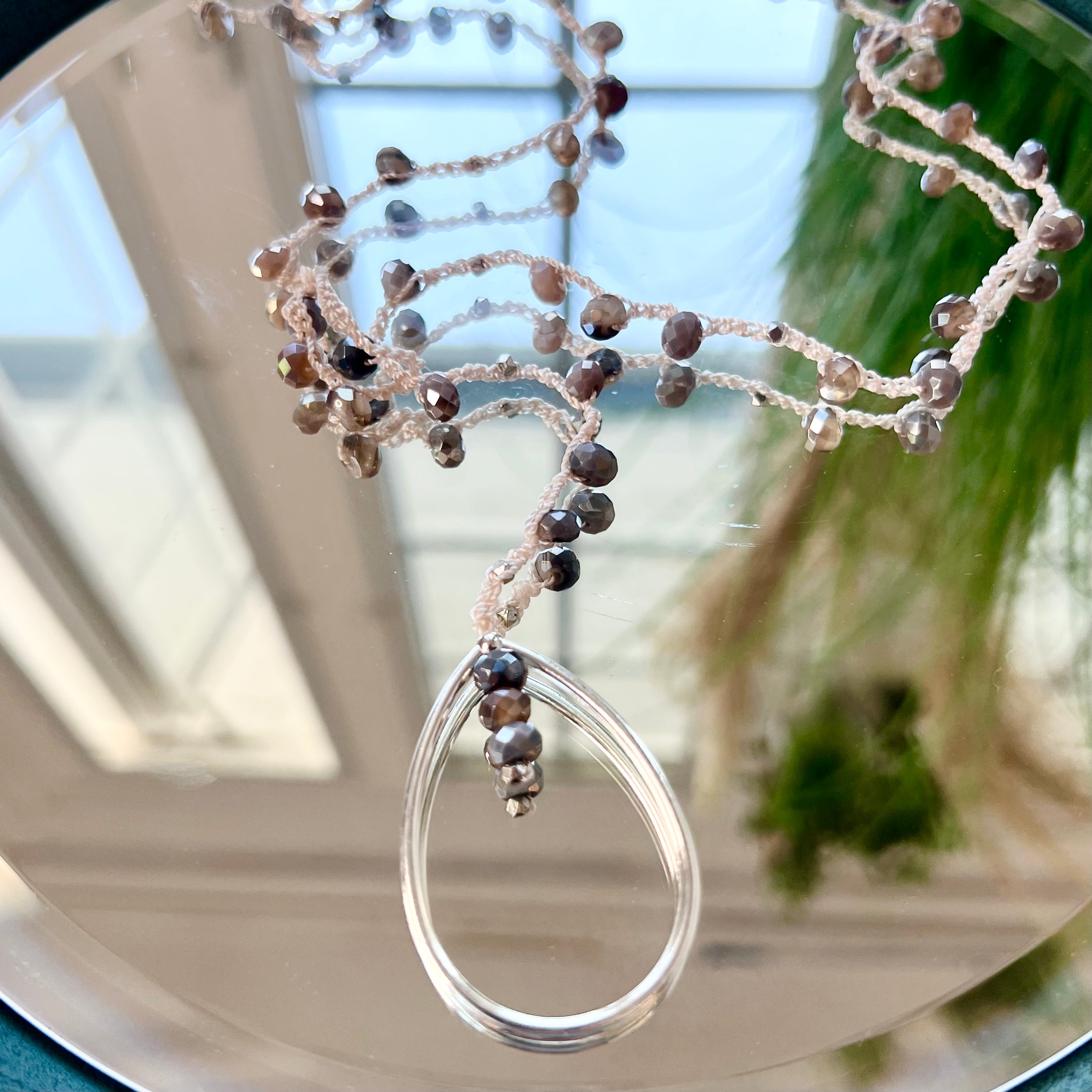 A crochet necklace with a teardrop-shaped pendant made of fossil agate stones, displayed in front of a mirror.