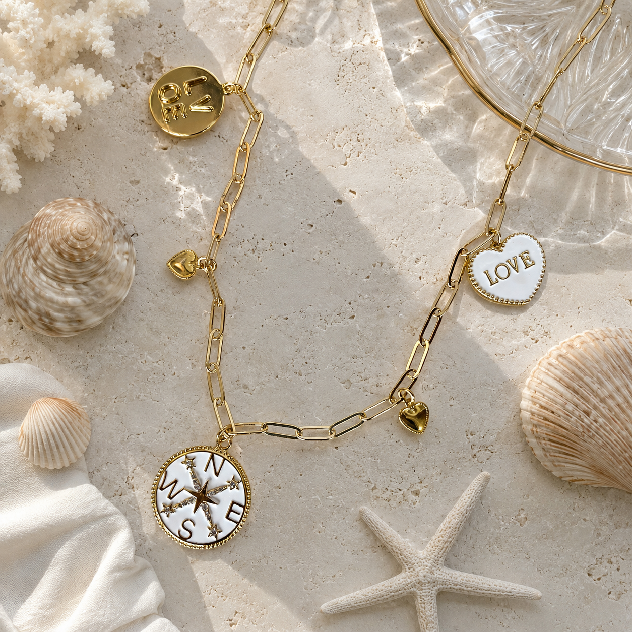 Gold bracelet with compass and 'LOVE' heart charms on a sandy surface with shells and starfish.