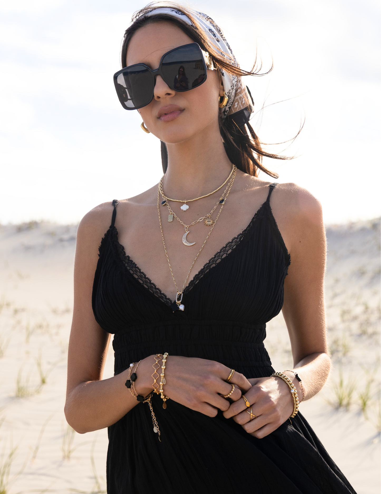 Woman in a black dress, sunglasses, the Payton Luna Star Necklace, customizable beaded choker in gold with a mother of pearl clover, retro hoops, and clover link bracelet.