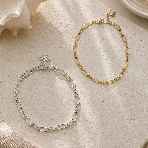 Two chain bracelets, one silver and one gold, on a textured beige surface with seashells and coral.