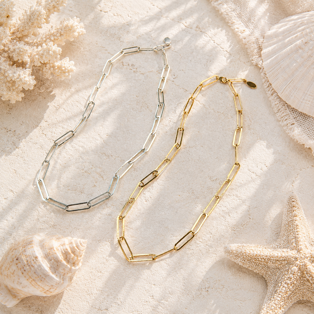 Two gold and silver chain necklaces on a sandy surface with shells and starfish.