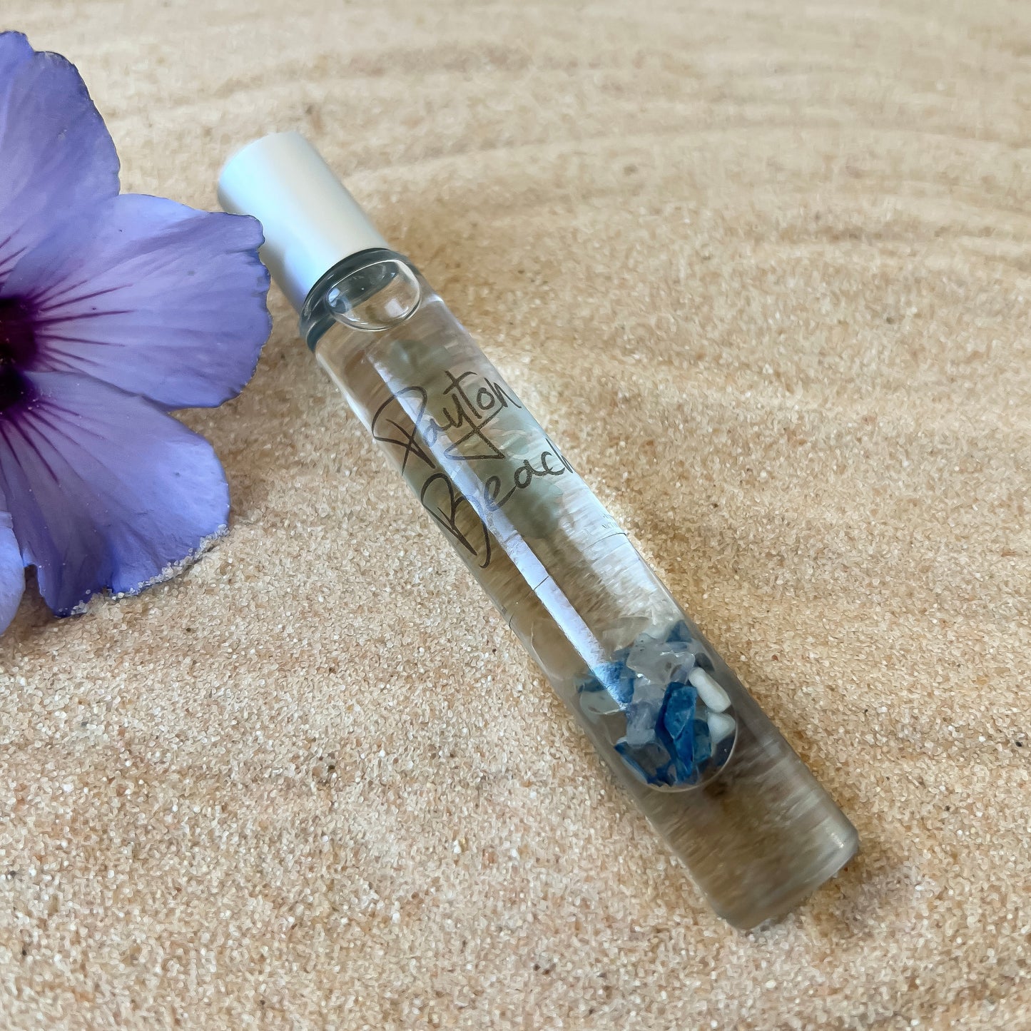 A clear aromatherapy roller with blue sea glass inside, placed on a sandy surface next to a purple flower.