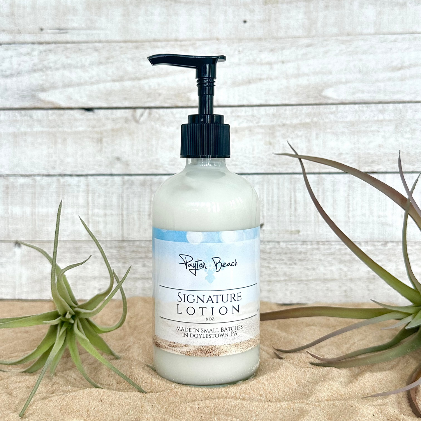 Payton Beach signature lotion, with hints of vanilla and coconut in a pump bottle  on a wooden surface with plants.