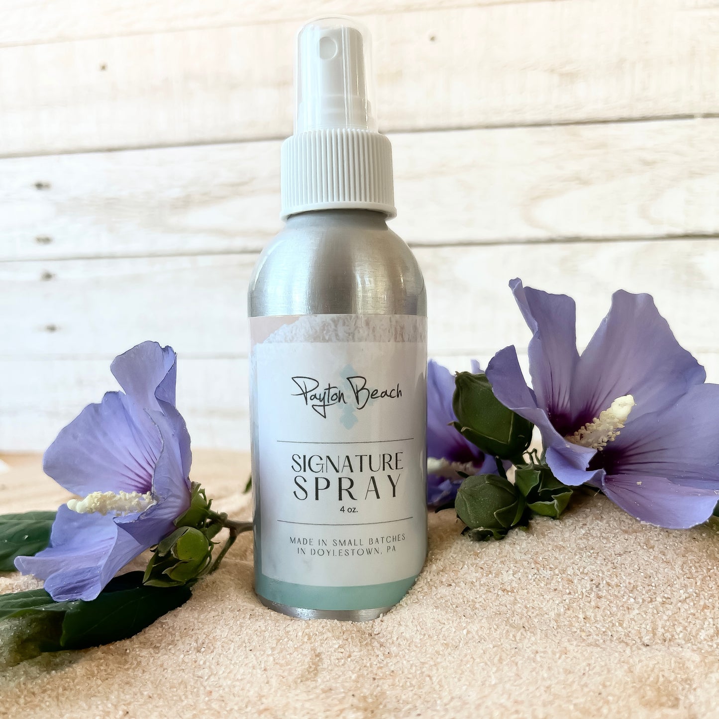A bottle of 'Payton Beach Signature Spray' with a spray pump, placed on a surface with sand and accompanied by two purple flowers.