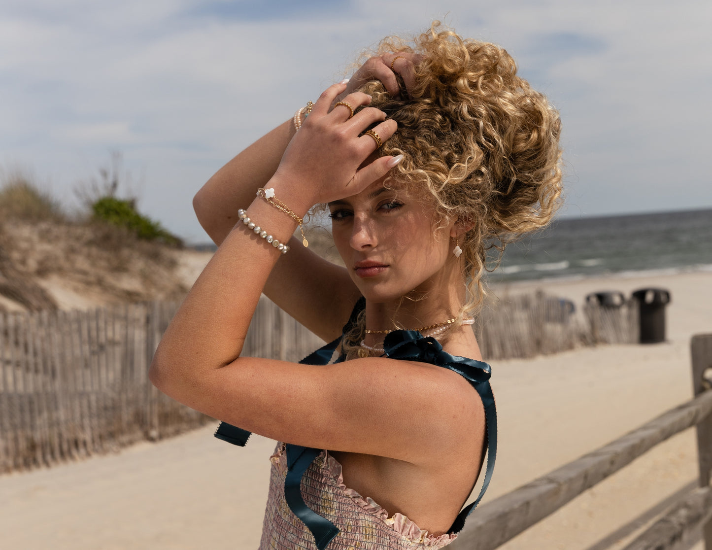 Woman with curly hair on a beach styled with Payton jewelry from head to toe. Including our island pearls, payton clovers, and custom designs from our charm bar, pa and nj's original biggest and best.
