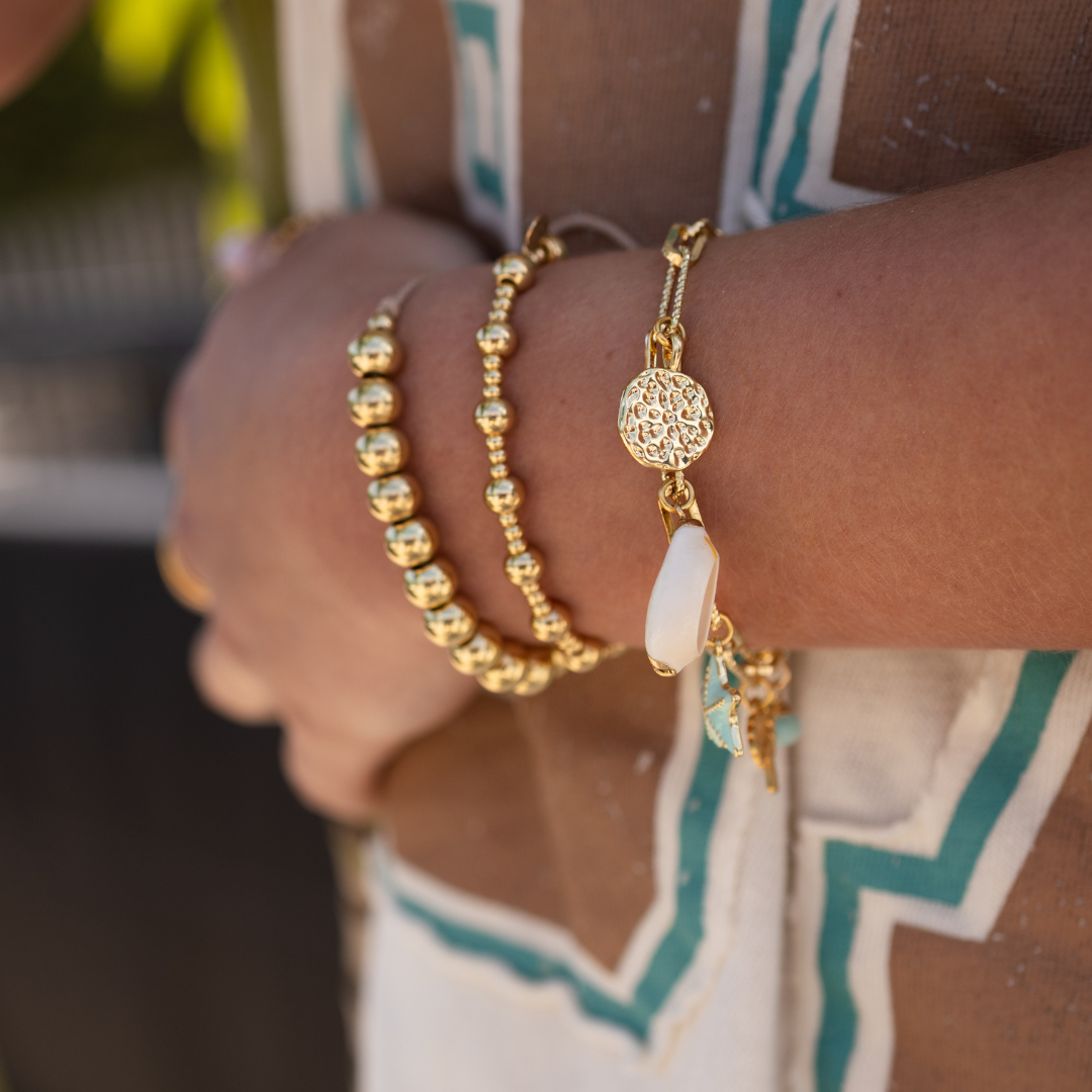 Close-up of a person's wrist wearing a gold charm bracelet, and two gold beaded bracelets with a blurred background