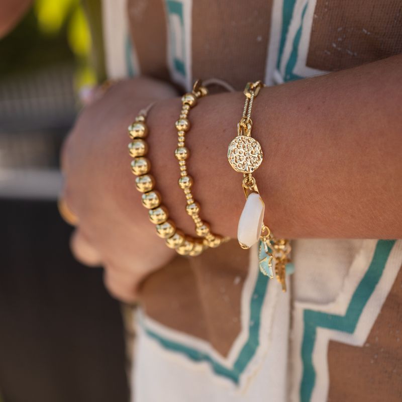 Close-up of a person's wrist wearing a gold charm bracelet, and two gold beaded bracelets with a blurred background