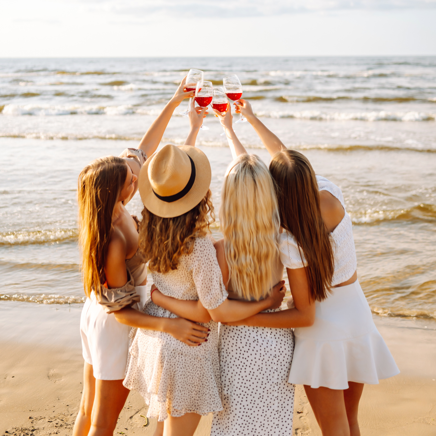 Four women on a beach, raising glasses together, with a scenic ocean view. Celebrate the special moments with Payton Jewelry at the original biggest and best charm bar in pa and nj.