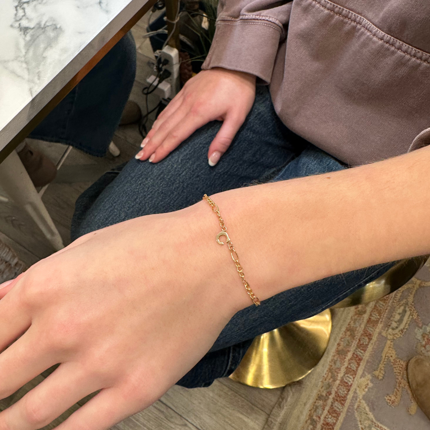 Close-up of a person's wrist wearing a gold permanent bracelet with a letter "c" connector charm.