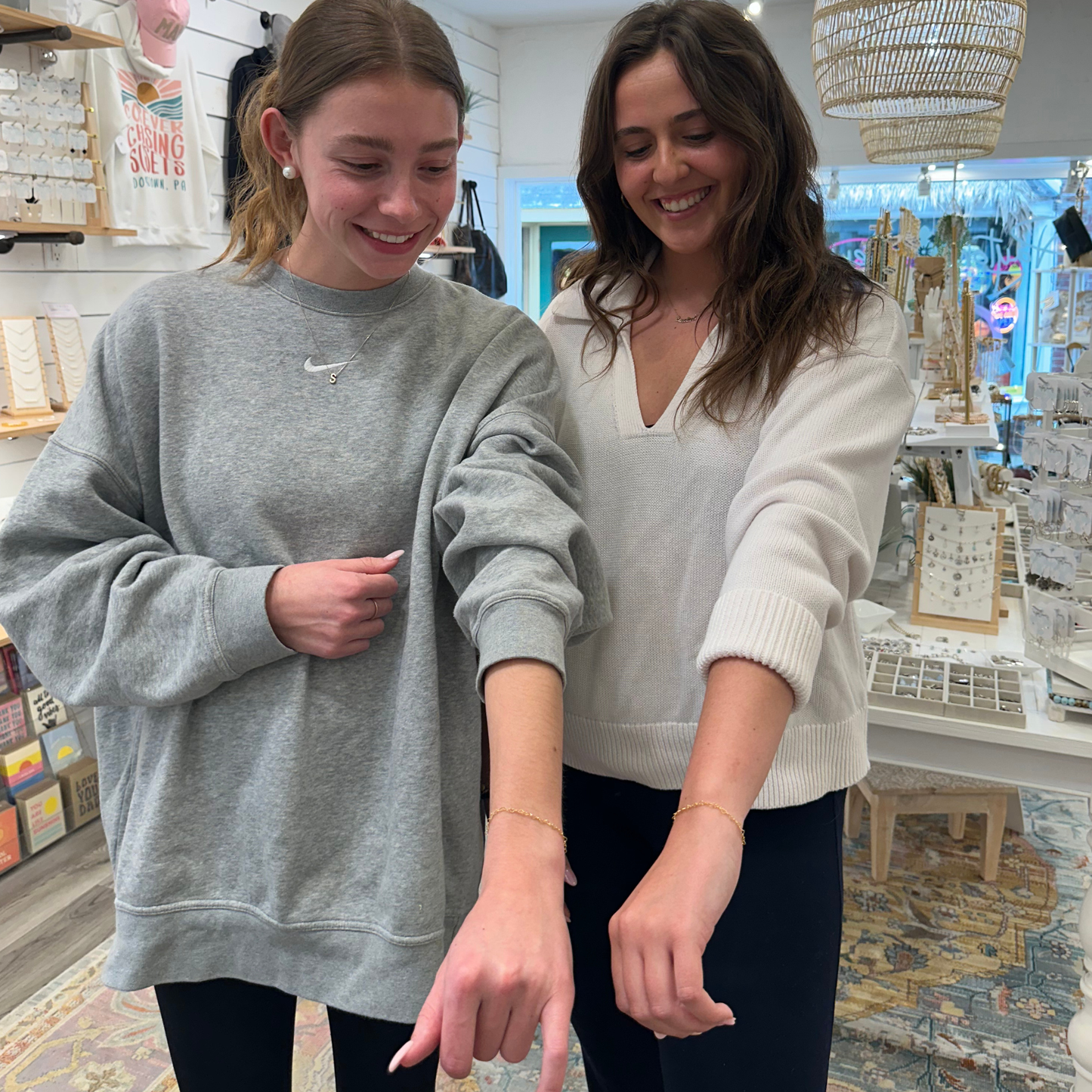 Two women standing in a store, showing off their matching permanent bracelets in gold.