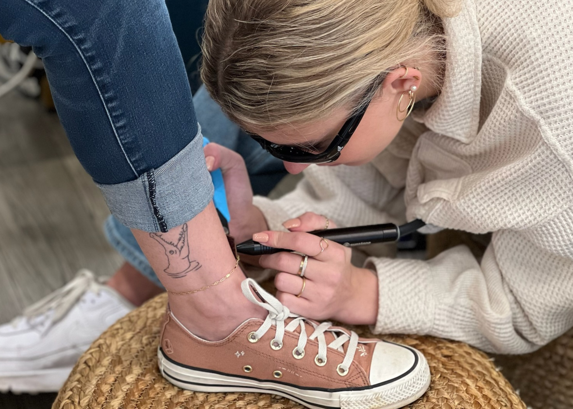 Person applying permanent jewelry on another person's ankle with a brown sneaker.