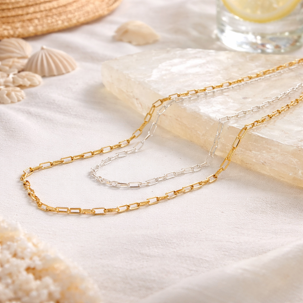 Multi layer chain in gold and silver on a sandy surface with seashells and a glass of water in the background.