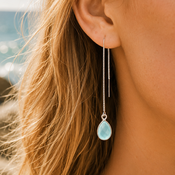 A close-up of a woman's ear wearing a long silver dangle earring with a light blue teardrop-shaped stone, set against a softly blurred outdoor background.