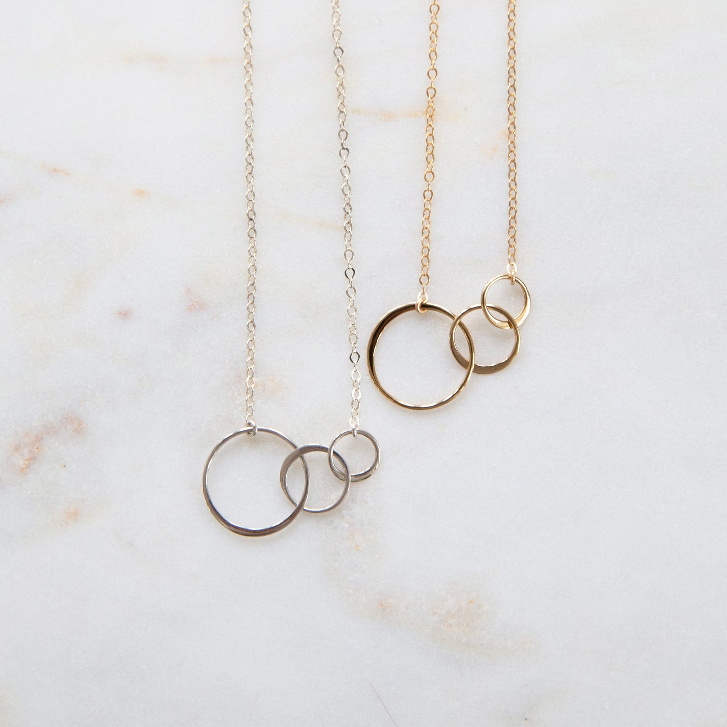 Two necklaces, one plated in gold and the other in silver, each with a series of three interlocking circles on a marble surface.