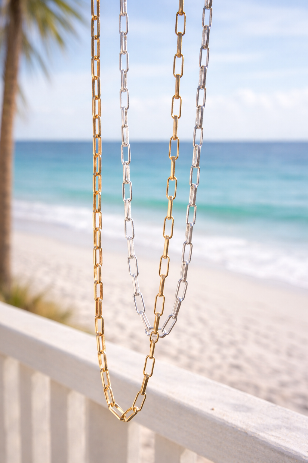 Two chains, one gold one silver,  on a white railing with a beach background