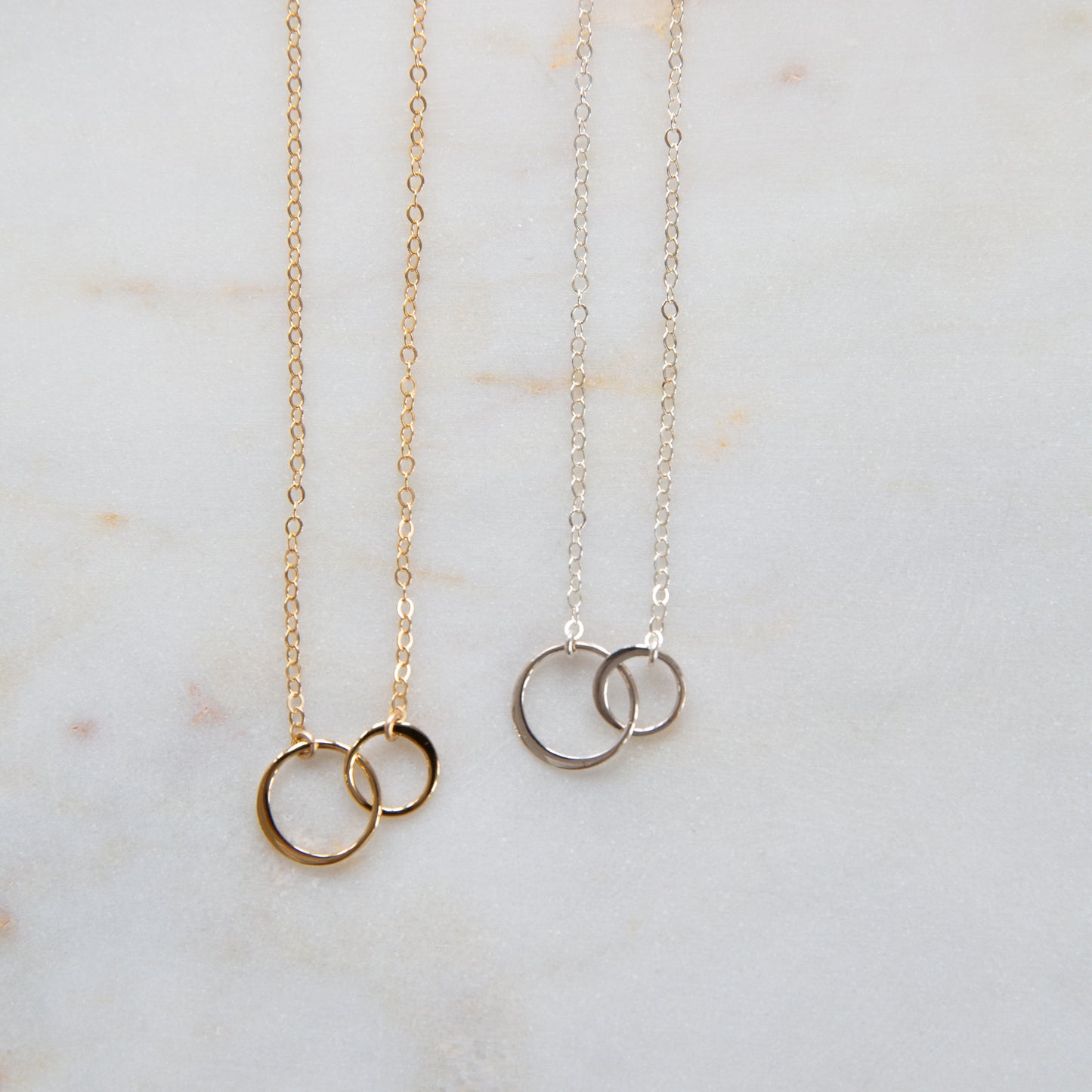 Two necklaces, one with gold chains and circles, the other with silver chains and circles, displayed on a marble surface.