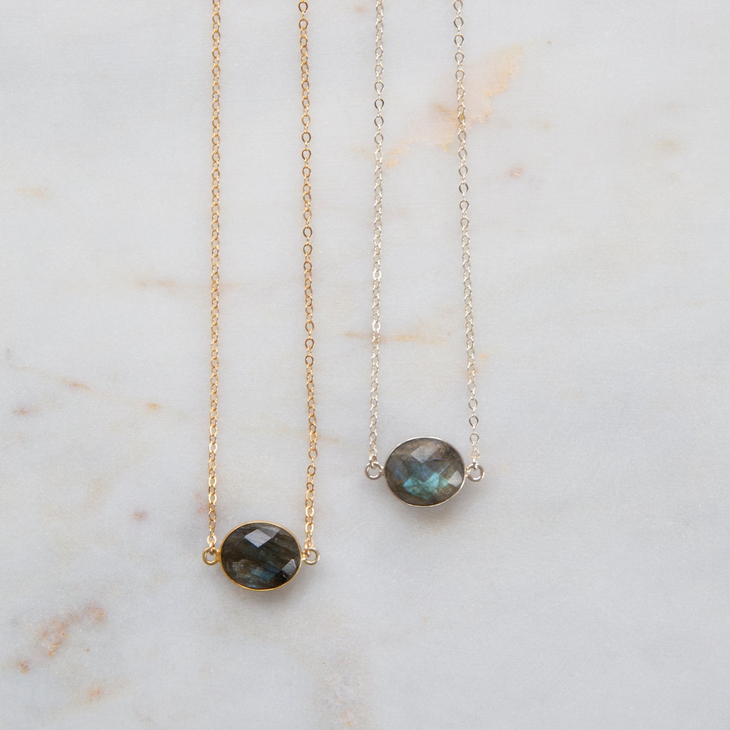 Two necklaces, one with a silver chain and a gold bezel holding a labradorite stone, and another with a gold chain and a gold bezel holding a labradorite stone, placed side by side on a marble surface.