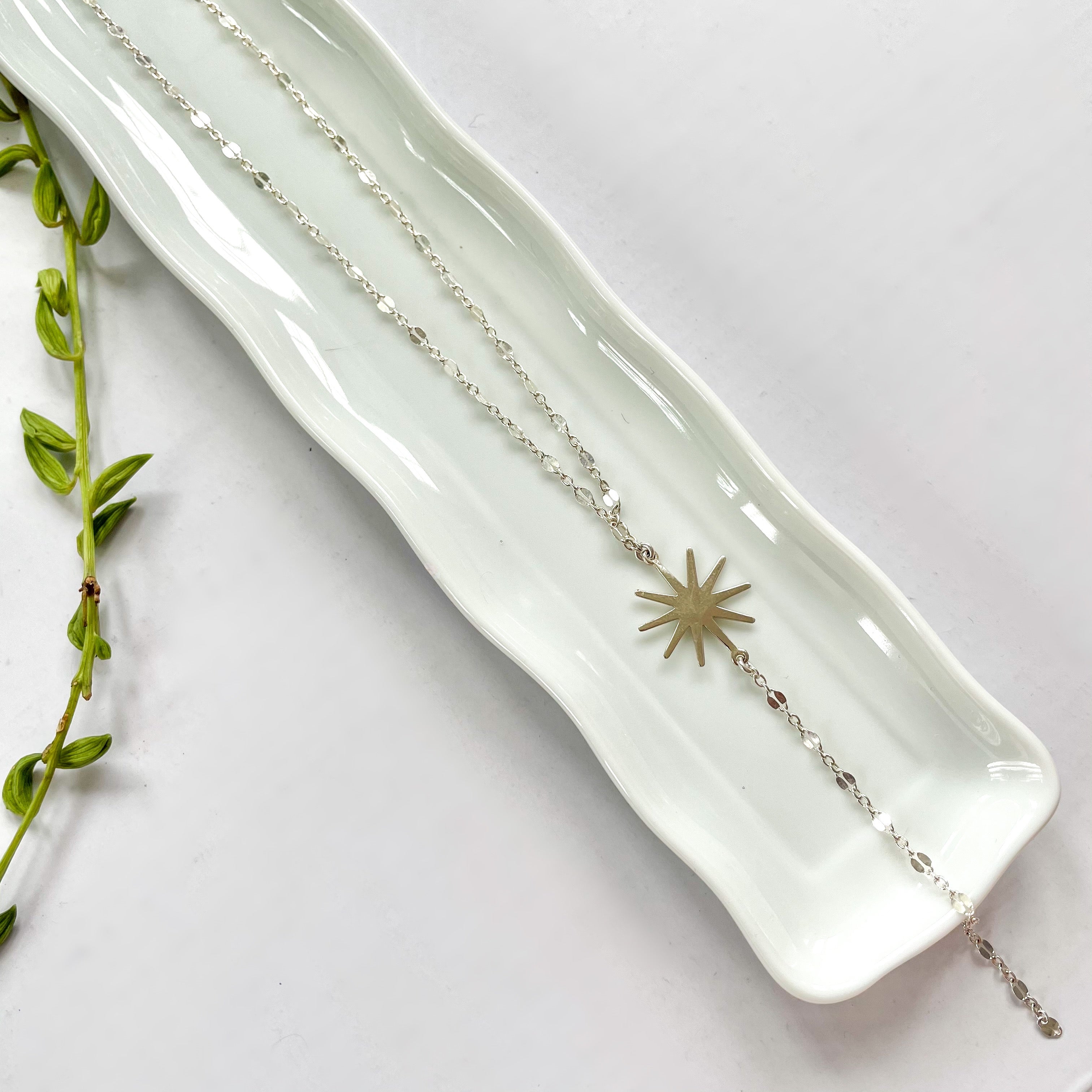 A delicate necklace with a metallic chain and a star-shaped pendant, displayed on a white plate with a green plant to the side.
