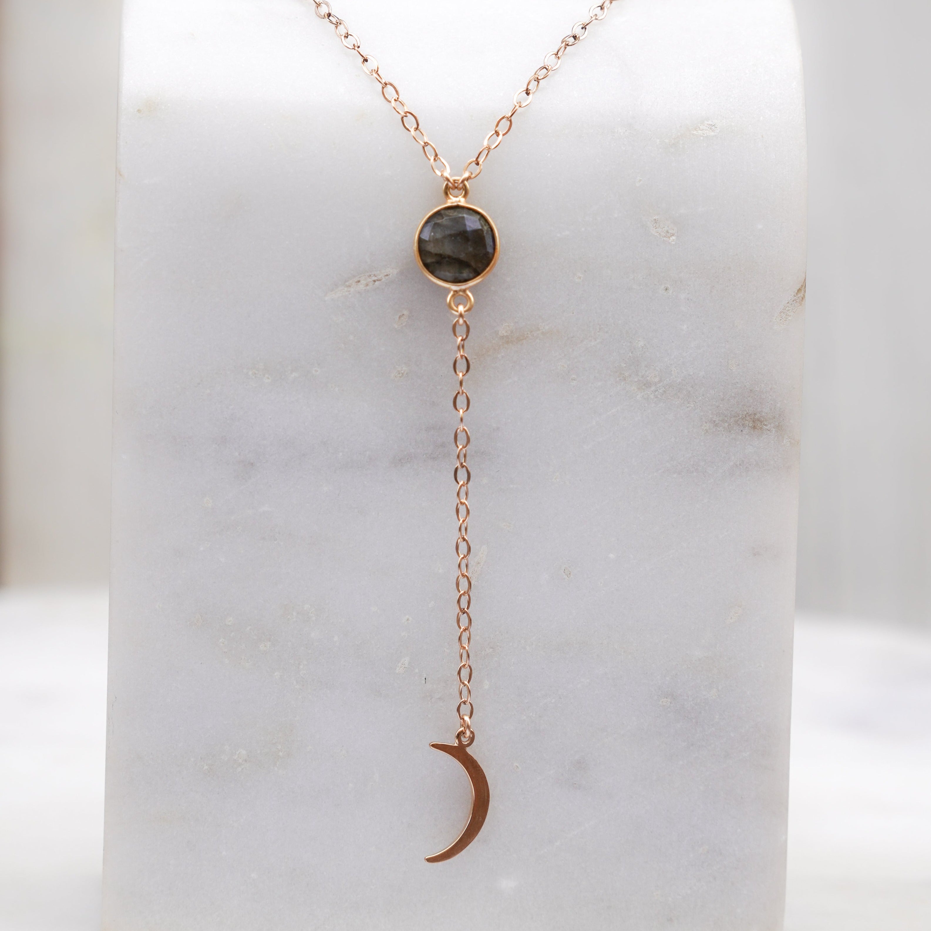 A Labradorite gem drop necklace with a crescent moon charm, displayed on a marble surface. The necklace is gold-filled, with the chain adjustable throughout.