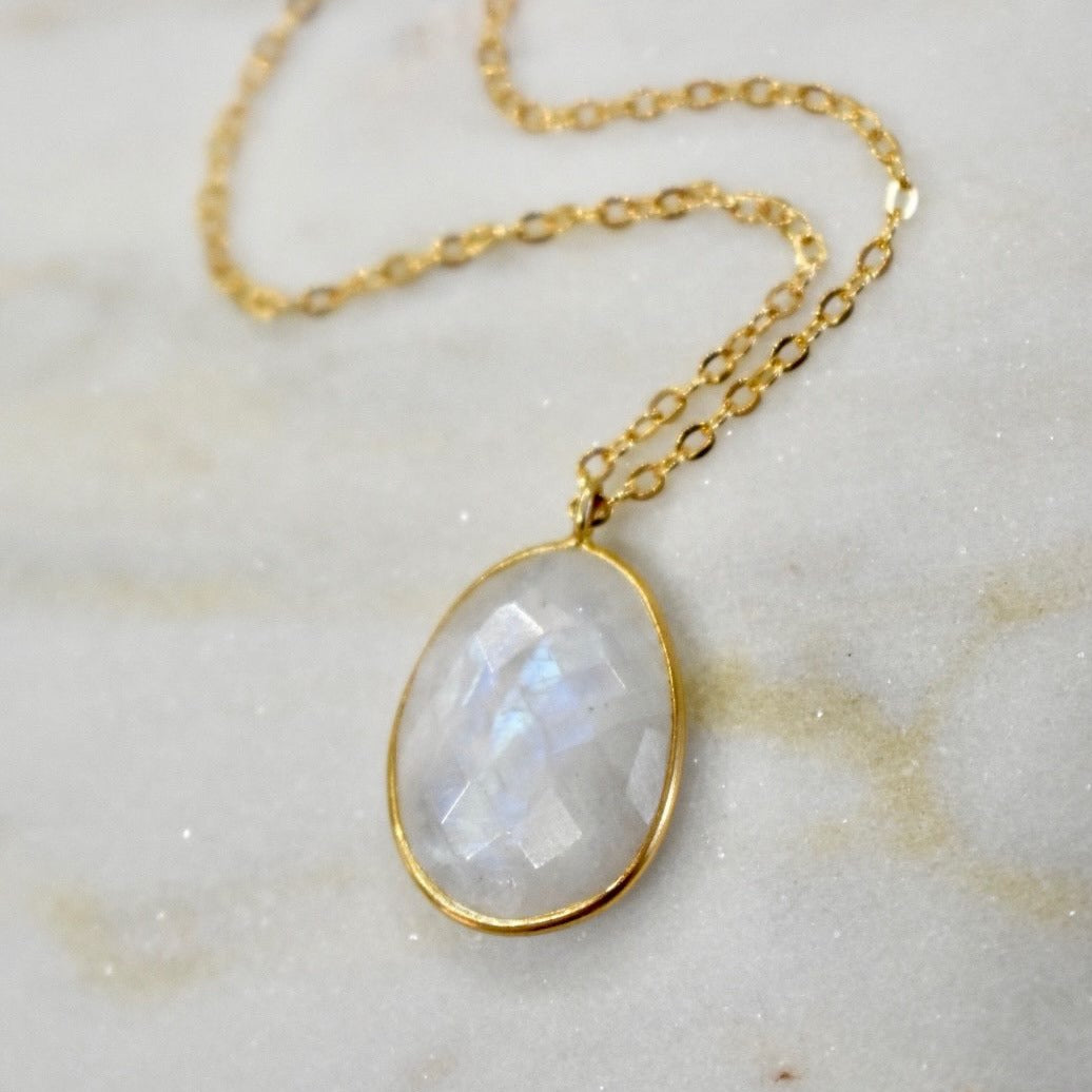 A gold chain necklace with a bezel setting containing a large, faceted moonstone pendant, displayed on a marble surface.
