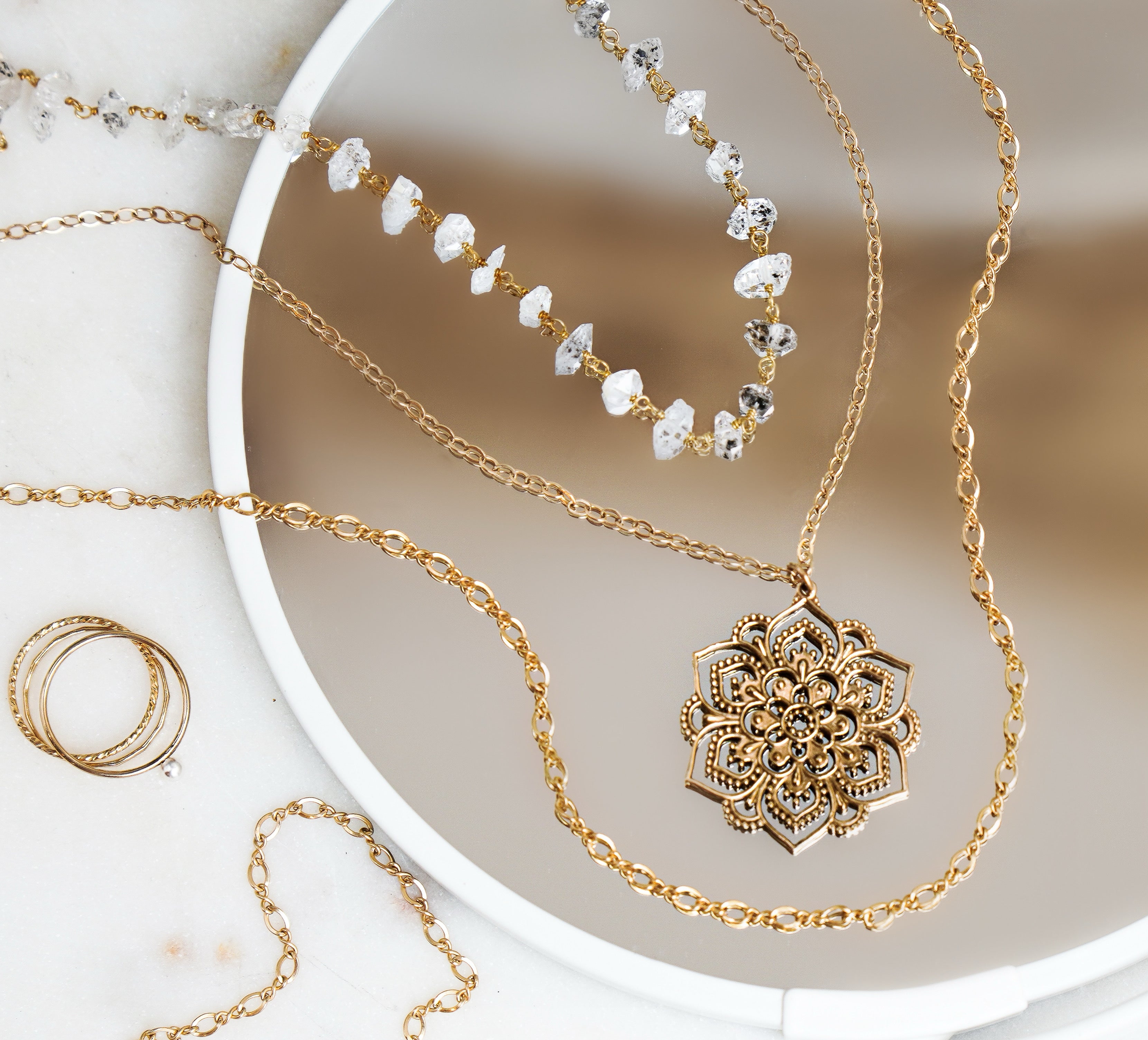 A gold fill mandala necklace displayed among other delicate jewelry pieces, placed on a white surface.