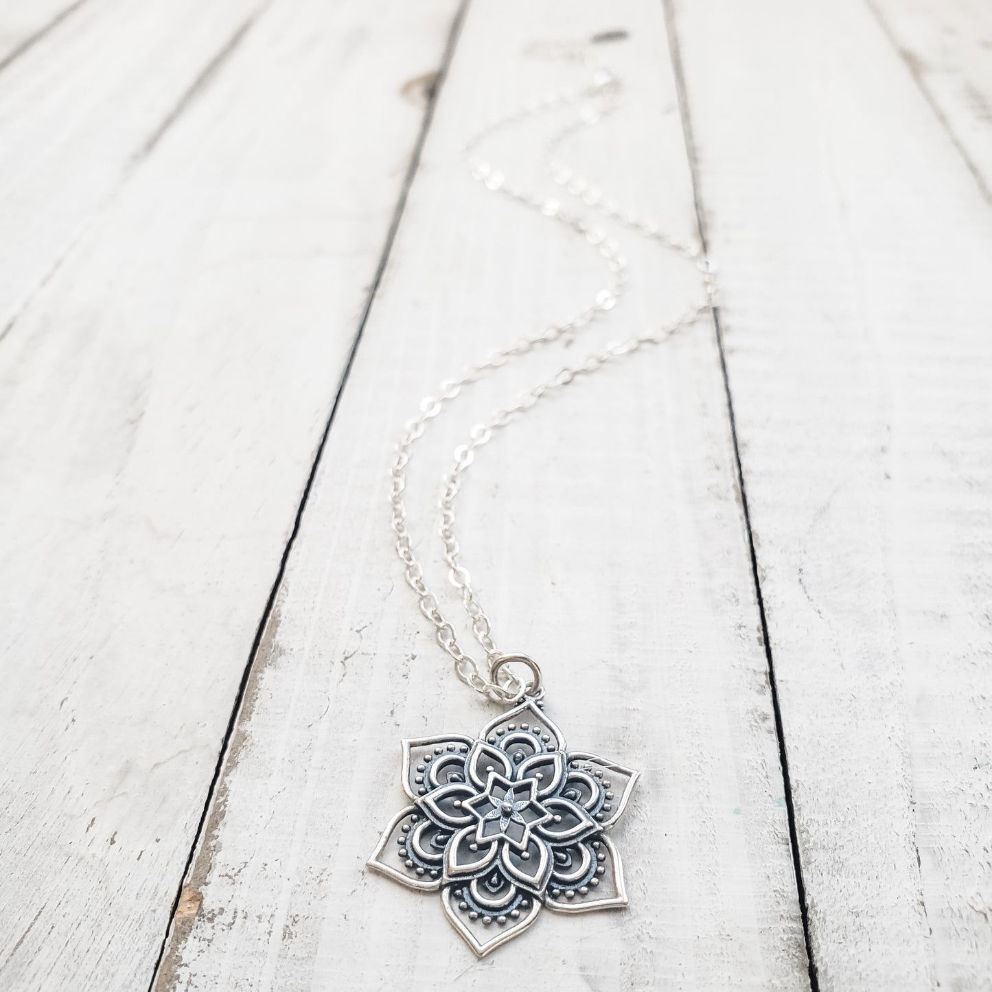A silver necklace with a floral mandala pendant laid out on a wooden surface.